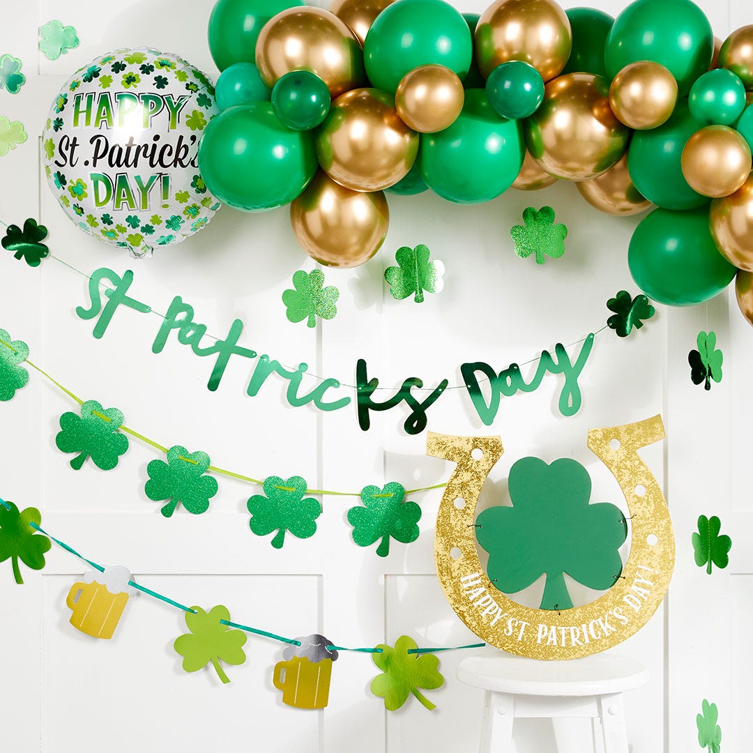 A green 'Happy St Patrick's Day' letter banner hanging on a wall with a green and gold balloon arch, and shamrock bunting